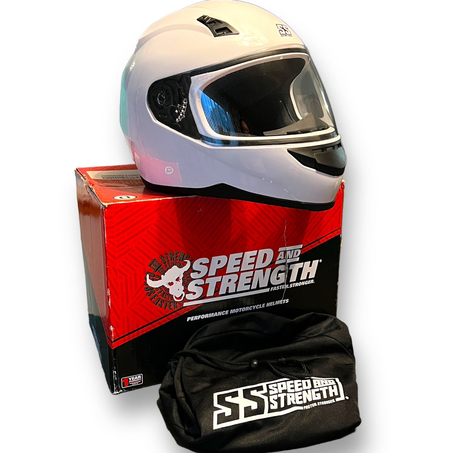 A gloss white full face motorcycle helmet with a clear visor, placed on top of its red and black box, with a black drawstring bag beside it.