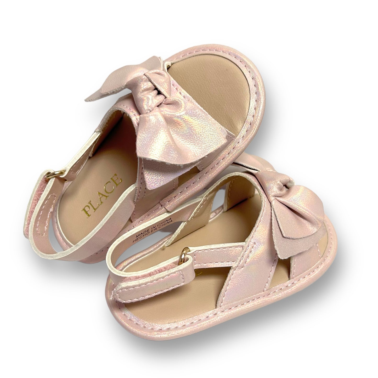 A pair of pink blush metallic shimmer sandals for children, with a bow detail on the ankle strap and a flat, platform sole.