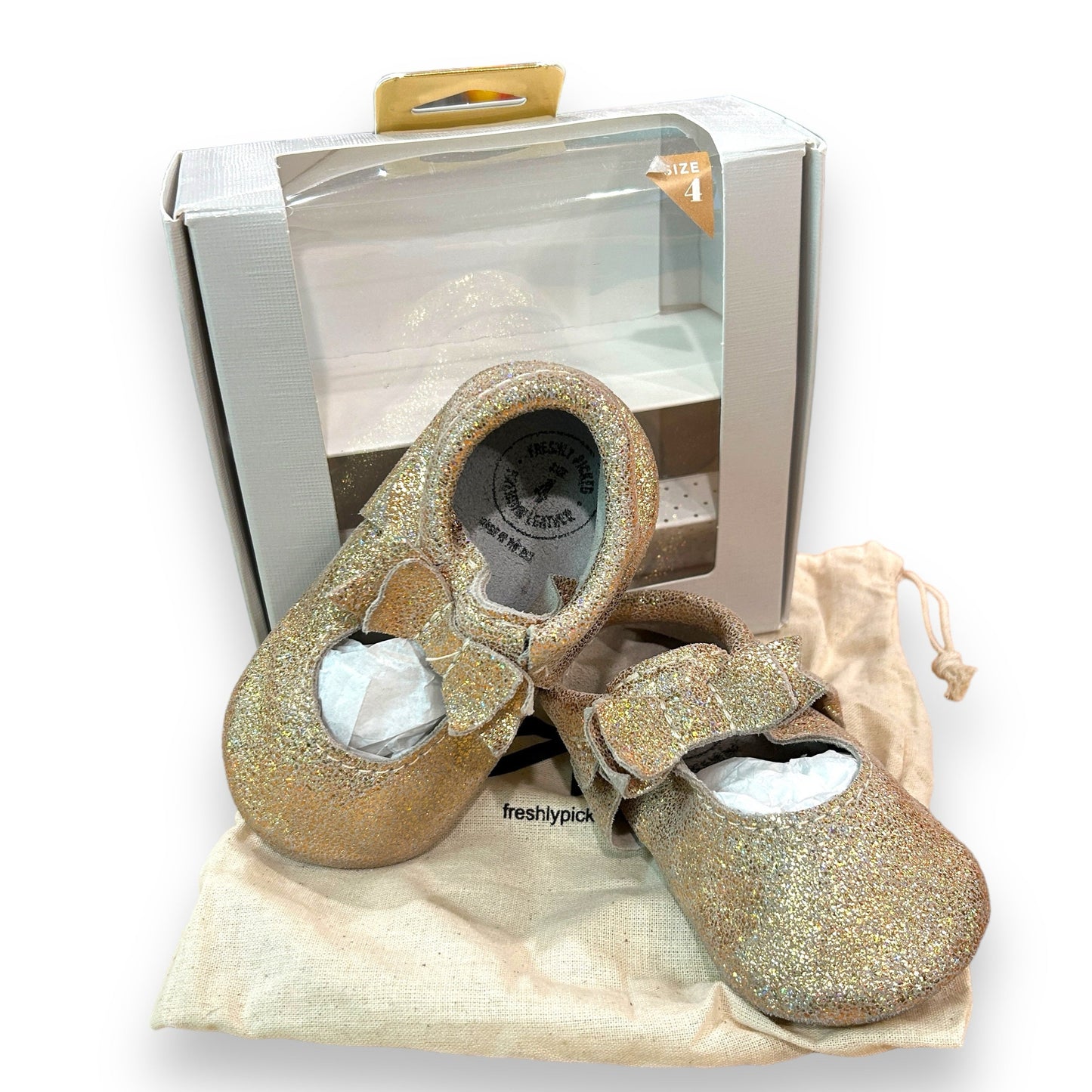 A pair of gold ballet flat bow mocc shoes for toddler girls, presented in an open box with a beige fabric lining.