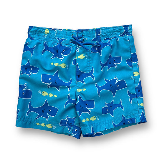A pair of blue swim trunks with a shark print design, featuring a drawstring waist and a brand label at the bottom.